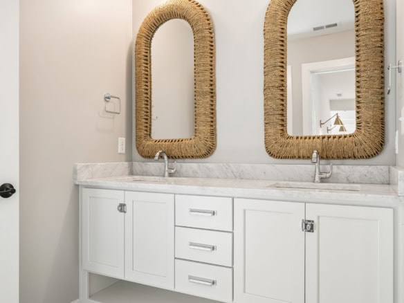 Bathroom vanity with two mirrors and wicker frames, white cabinetry, and marble countertop.