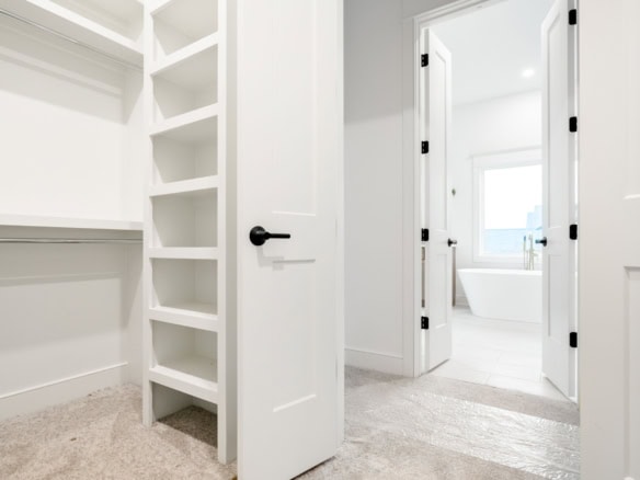 Image of a spacious, white walk-in closet with built-in shelves, leading to a bright bathroom with a.