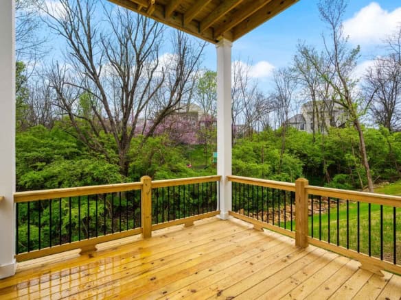 Wooden balcony overlooking lush green trees and a house in the distance.