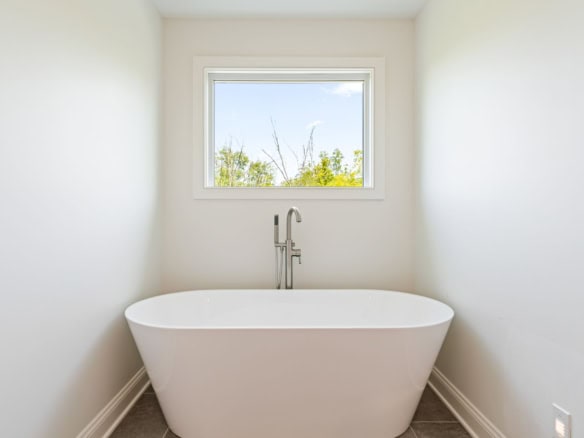 A bright, minimalist bathroom featuring a white freestanding bathtub beneath a window with a scenic.