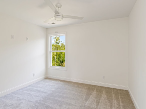 A bright, empty bedroom with white walls, a ceiling fan, and a window showing greenery outside.