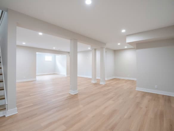 Interior view of a spacious, finished basement with white walls, wooden flooring, and multiple suppo.