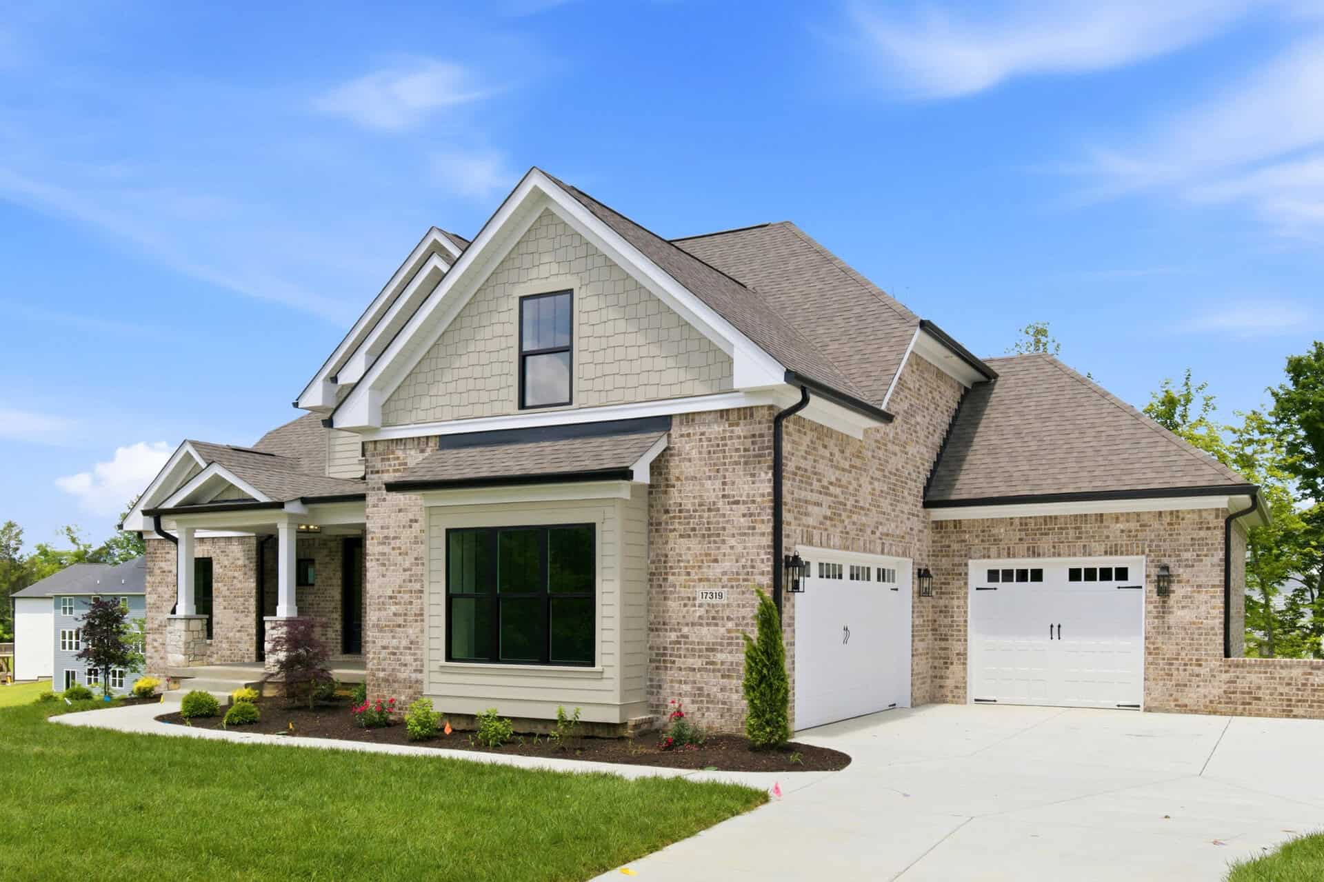 Front view of a modern brick suburban home with beige siding, large black-framed windows, and a two-car white garage under a clear blue sky.