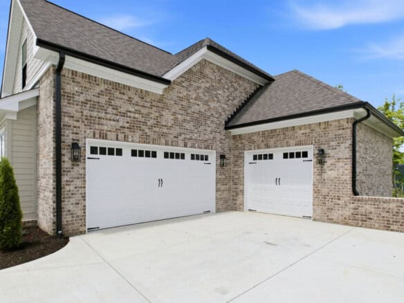 Front view of a brick house with two white carriage-style garage doors and a concrete driveway under a clear blue sky.