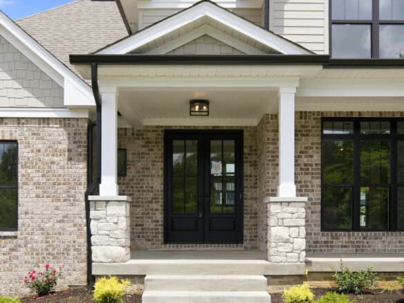 Front entrance of a brick house with white trim, black double doors, and a covered porch supported by stone columns.