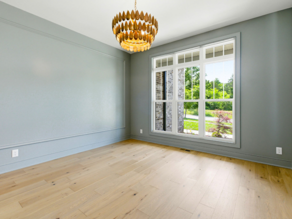 An empty light-filled room with gray walls, a large window revealing greenery outside, and a decorative wooden chandelier hanging from the ceiling.