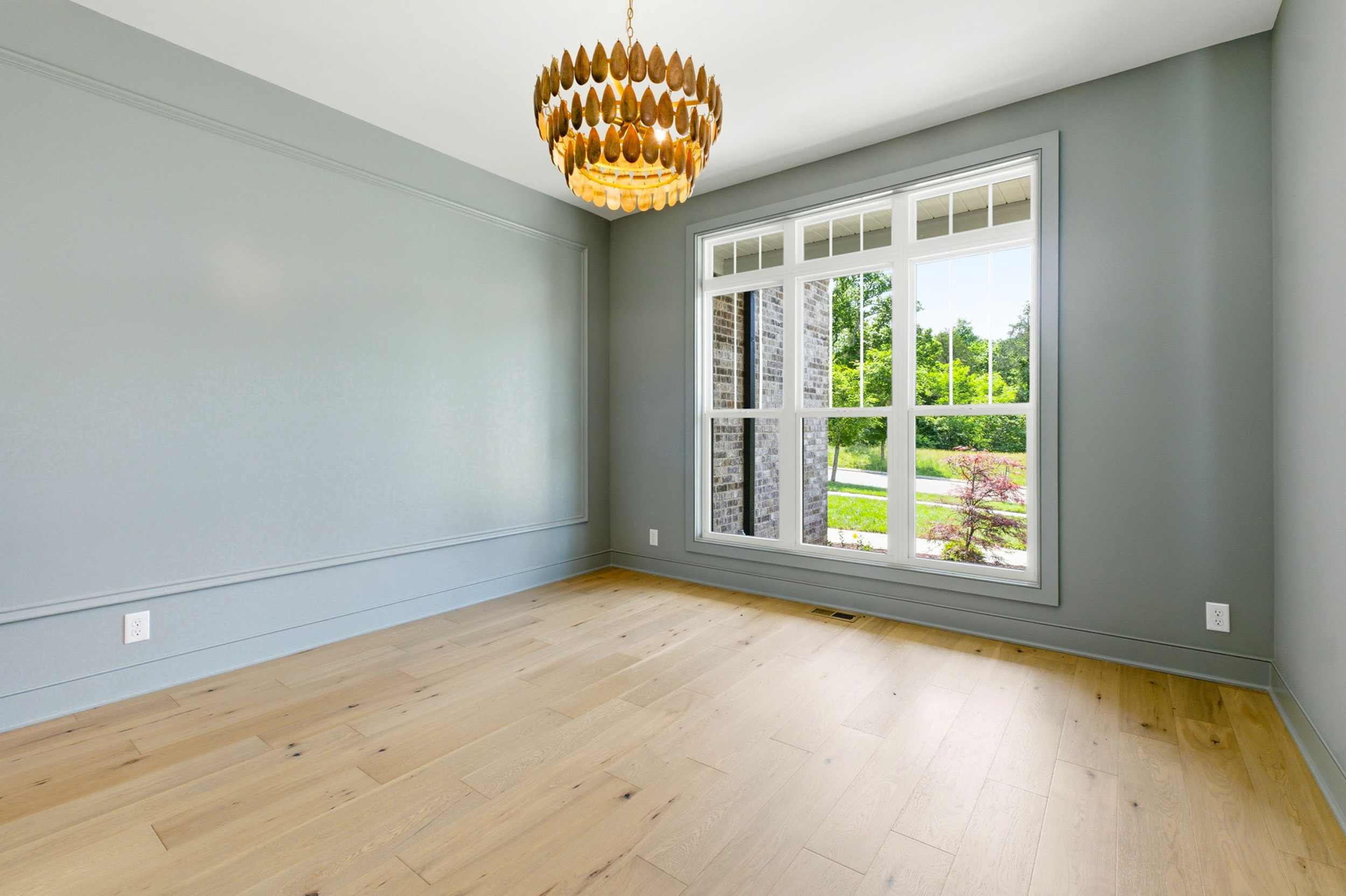 An empty light-filled room with gray walls, a large window revealing greenery outside, and a decorative wooden chandelier hanging from the ceiling.