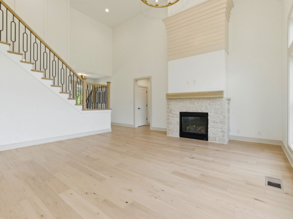 Bright living room with a white stone fireplace, light wood floors, and a staircase with decorative black railing.