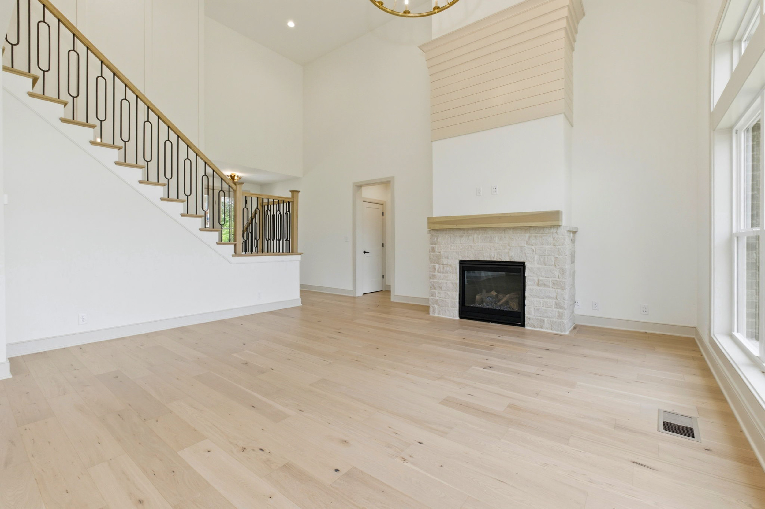Bright living room with a white stone fireplace, light wood floors, and a staircase with decorative black railing.