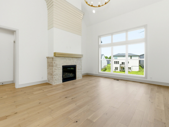 Bright empty living room with a stone fireplace, large windows, and light wood floors under a high white ceiling.