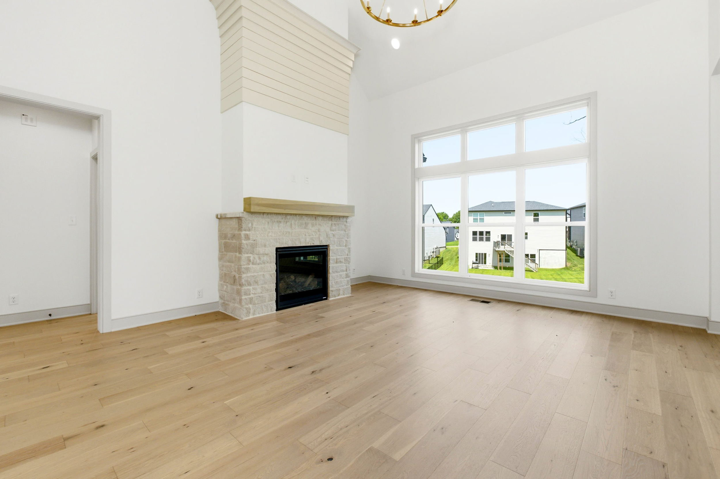 Bright empty living room with a stone fireplace, large windows, and light wood floors under a high white ceiling.