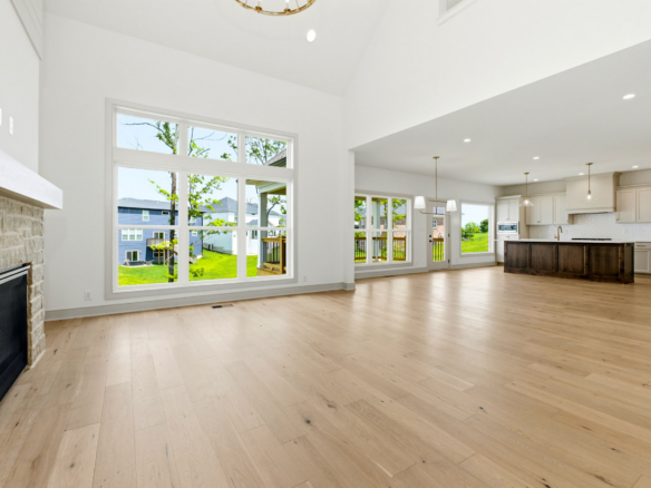 Open-concept living room with tall windows, light wood floors, stone fireplace on the left, and a modern kitchen island in the background.