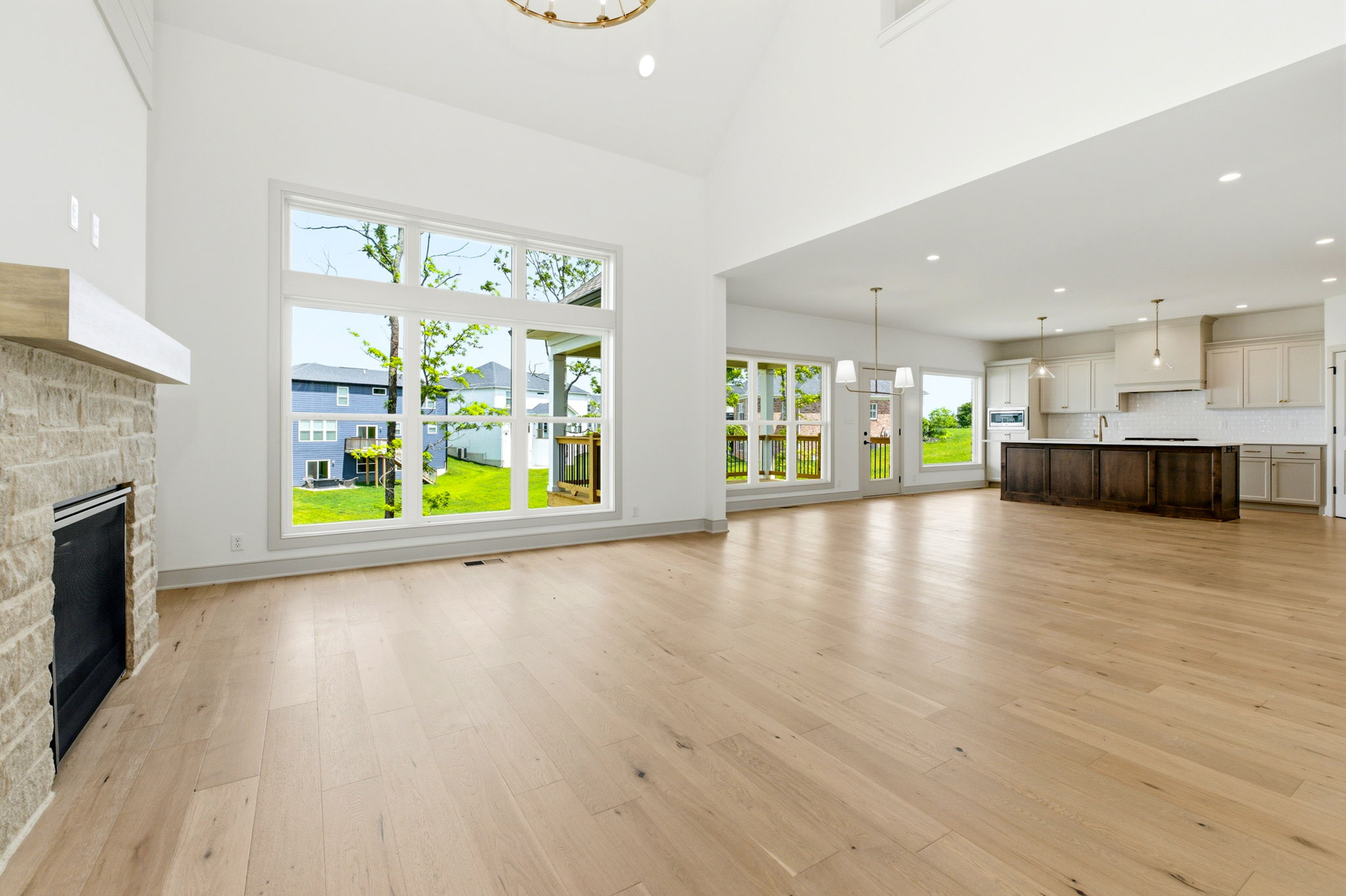 Open-concept living room with tall windows, light wood floors, stone fireplace on the left, and a modern kitchen island in the background.