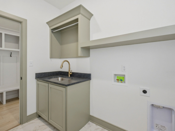 Utility kitchenette with gray cabinets, dark countertop, and a gold faucet, plus open wall cabinet and laundry hookups on the right wall.