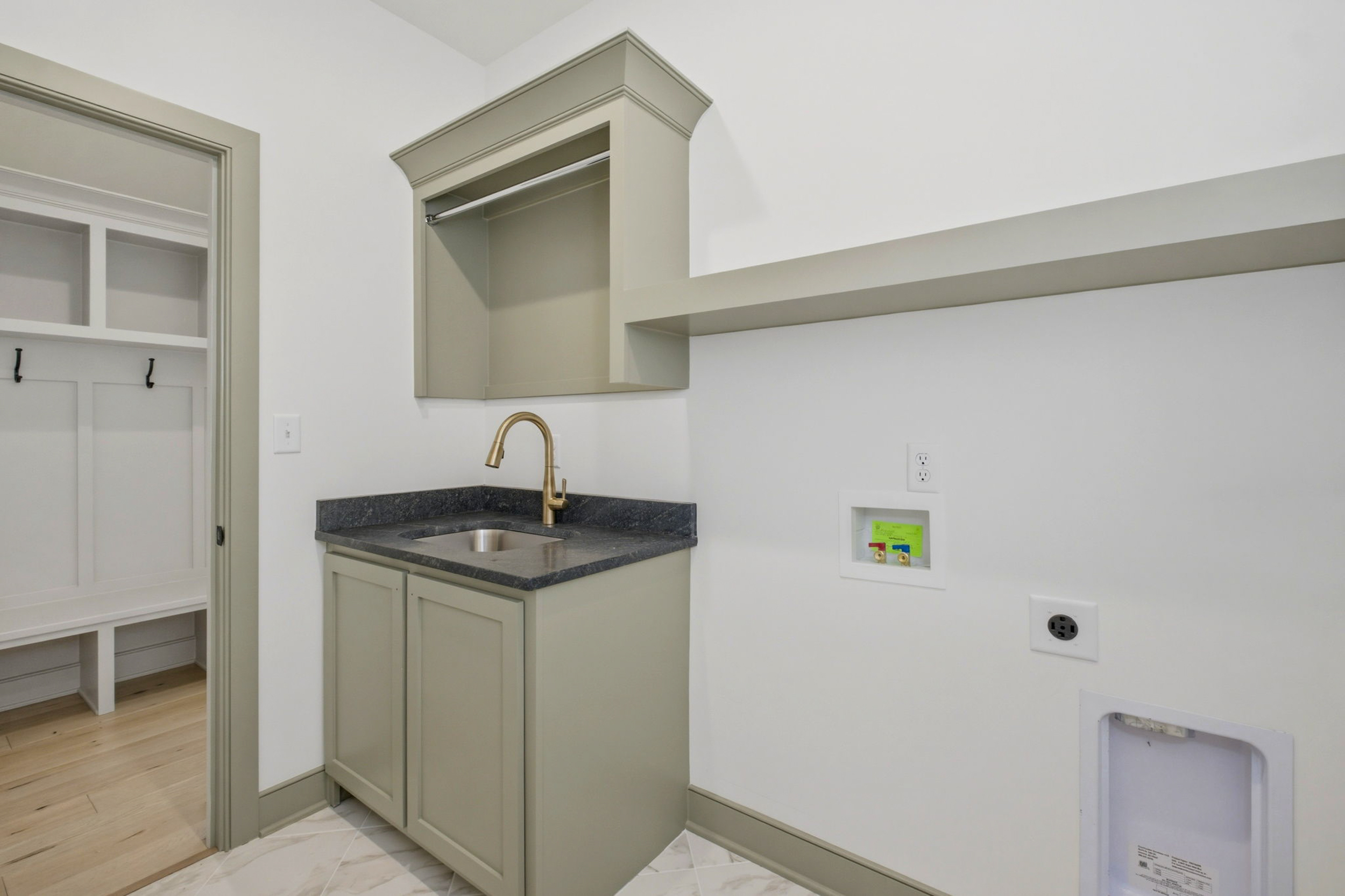 Utility kitchenette with gray cabinets, dark countertop, and a gold faucet, plus open wall cabinet and laundry hookups on the right wall.