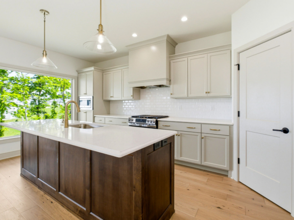 Bright kitchen with white cabinets and marble island, brass pendant lights, and a large window showing green trees outside.