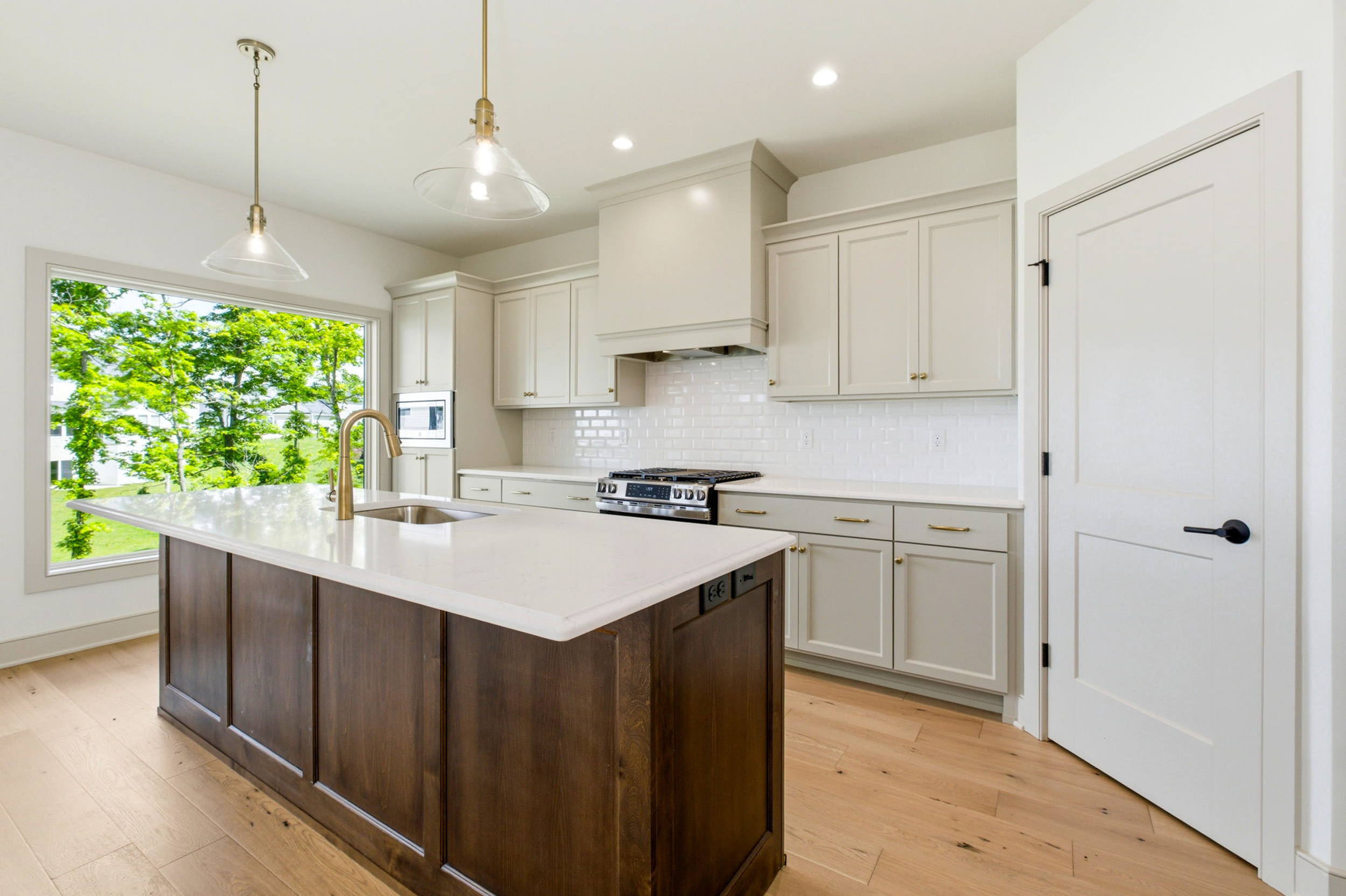Bright kitchen with white cabinets and marble island, brass pendant lights, and a large window showing green trees outside.