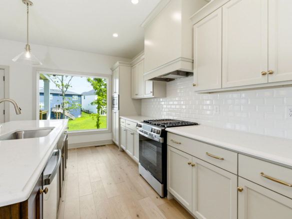 Bright white kitchen with an island, stainless steel stove, and a large window showing a green backyard.