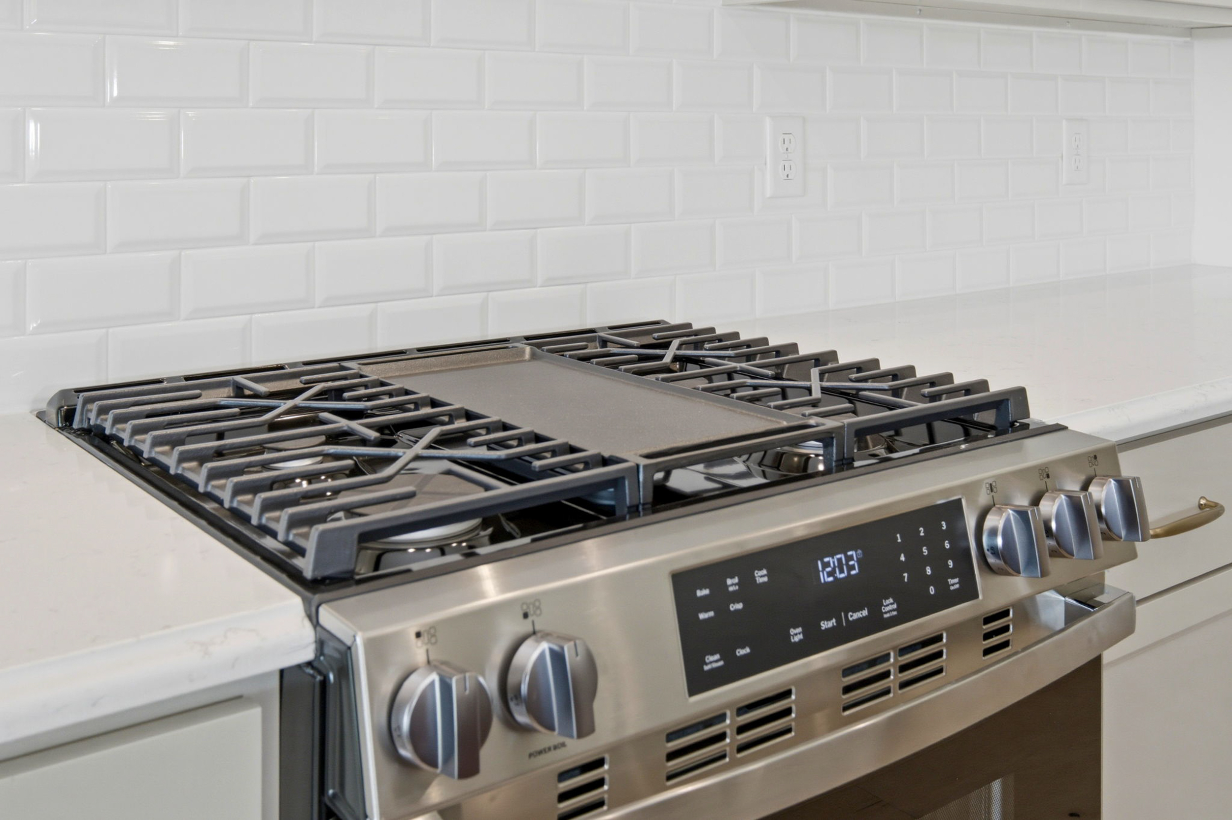 Stainless steel gas range with four burners and control panel against a white subway tile backsplash.