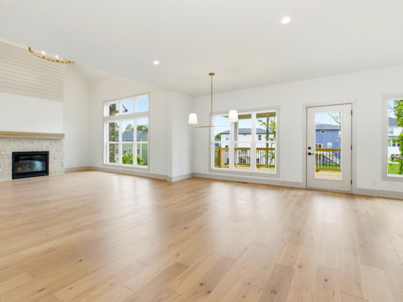 Bright, empty living room with a stone fireplace on the left, light hardwood floors, and large windows letting in natural light. A glass door leads to an outdoor deck.