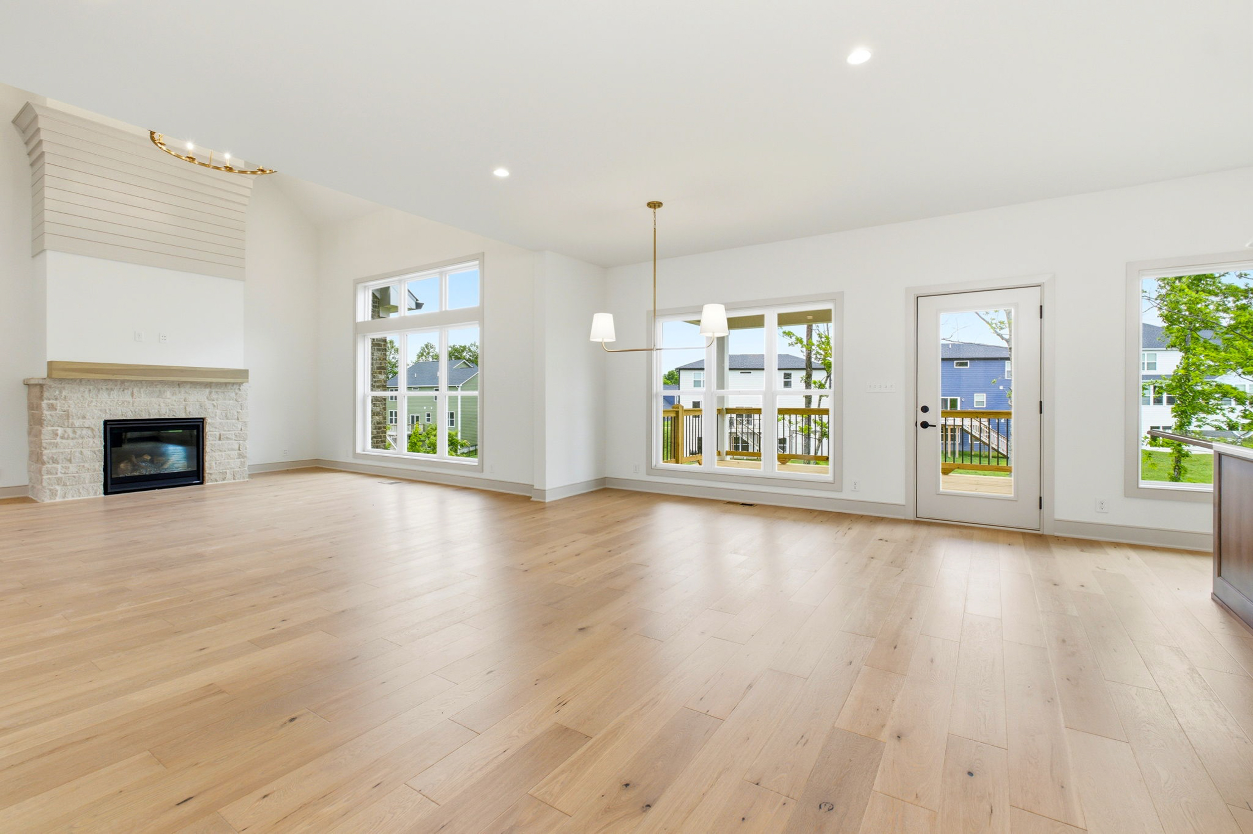 Bright, empty living room with a stone fireplace on the left, light hardwood floors, and large windows letting in natural light. A glass door leads to an outdoor deck.