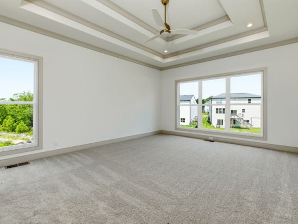 Bright, empty room with a tray ceiling, ceiling fan, beige trim, and large windows showing a suburban neighborhood