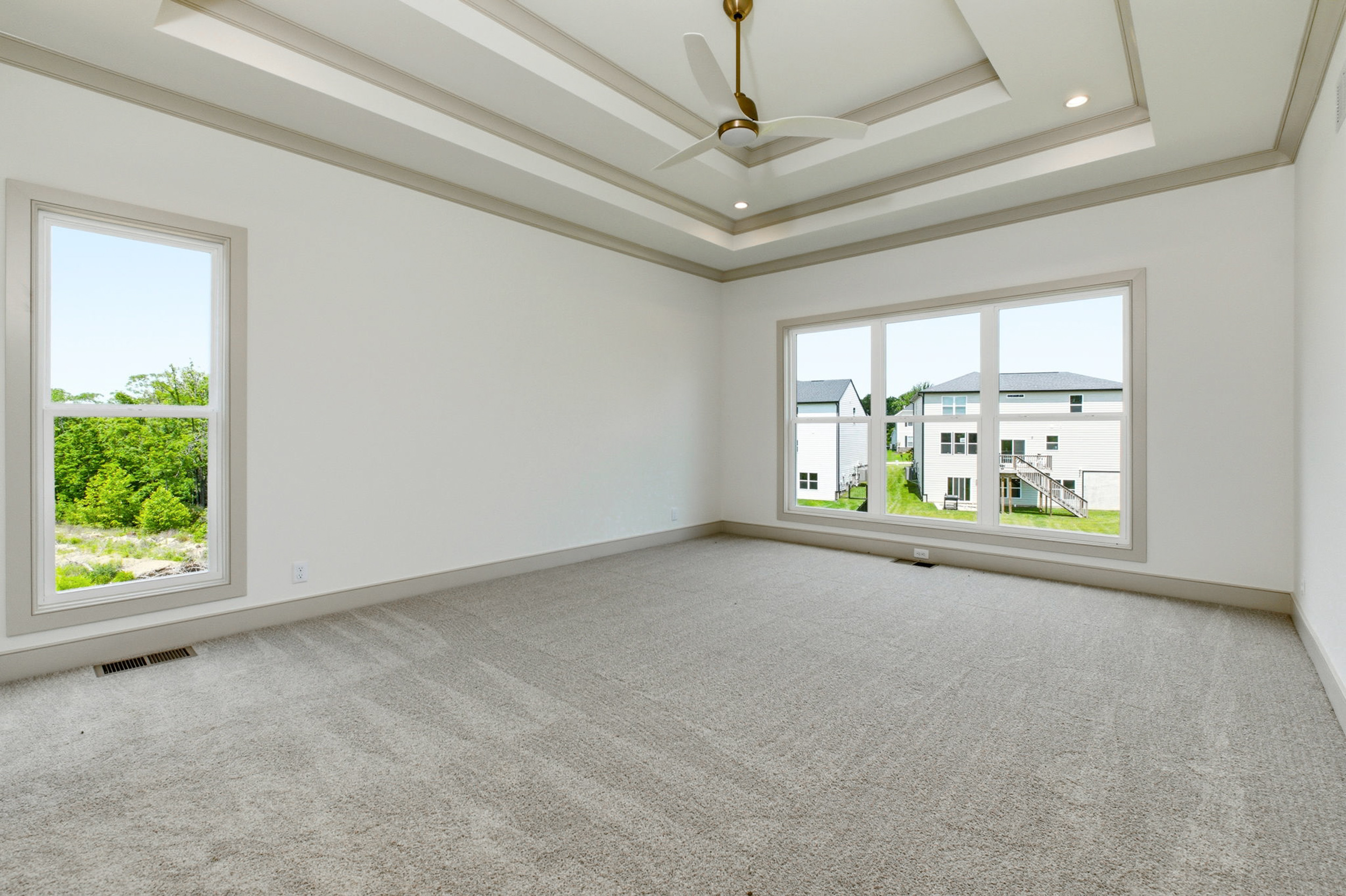 Bright, empty room with a tray ceiling, ceiling fan, beige trim, and large windows showing a suburban neighborhood