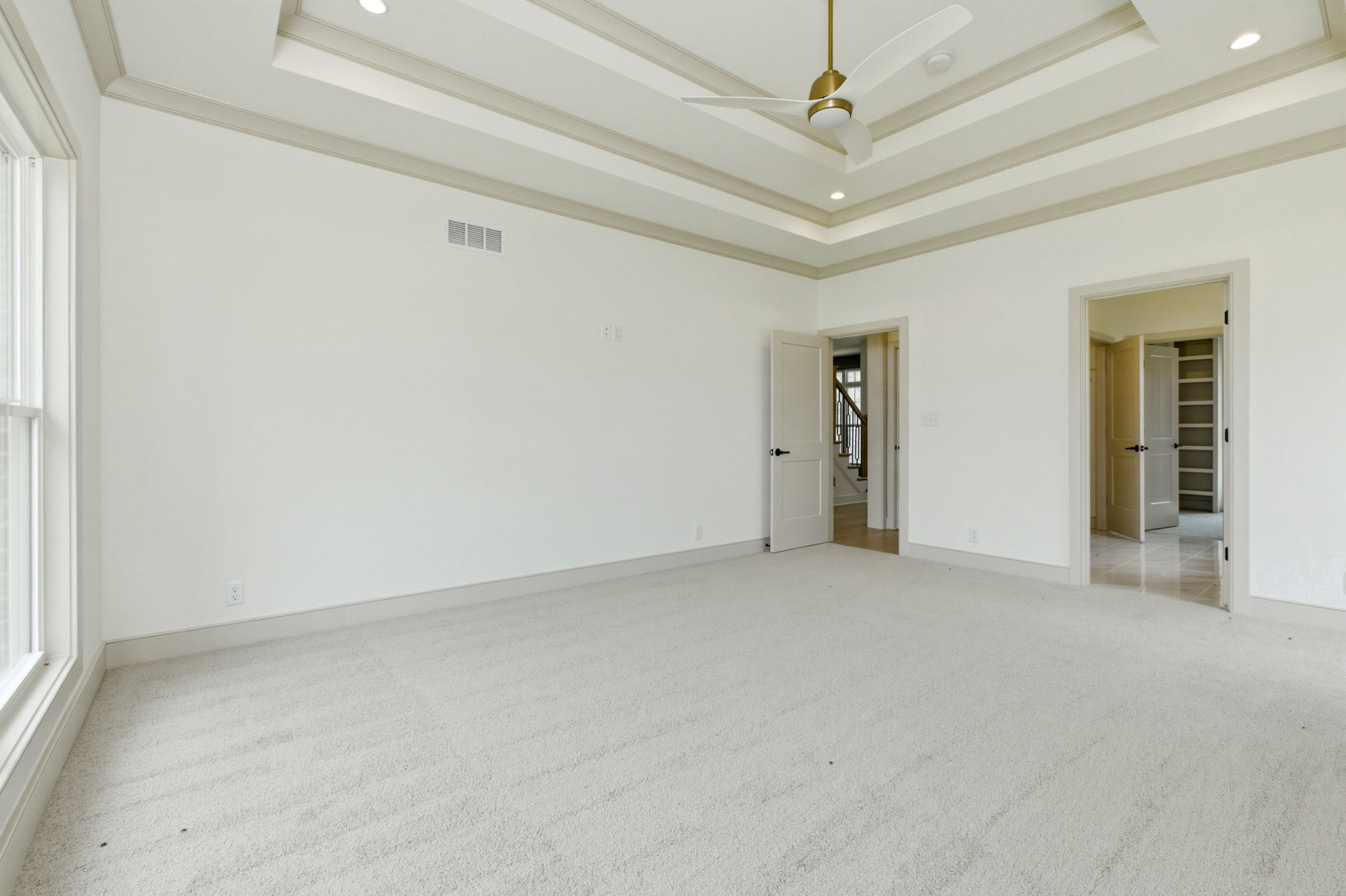 Bright empty living room with white walls, carpet, and a tray ceiling with a ceiling fan; doorway to another room visible on the right.