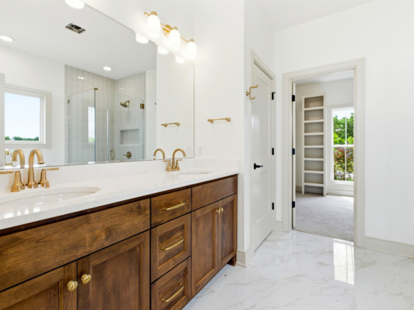 Double vanity with dark wood cabinets and gold faucets in a bright white bathroom; glass shower and open doorway to a room with built-in shelves.