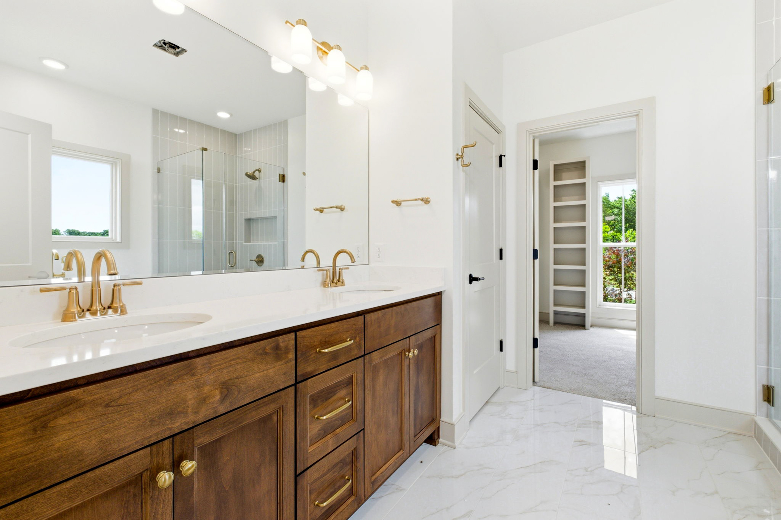 Double vanity with dark wood cabinets and gold faucets in a bright white bathroom; glass shower and open doorway to a room with built-in shelves.