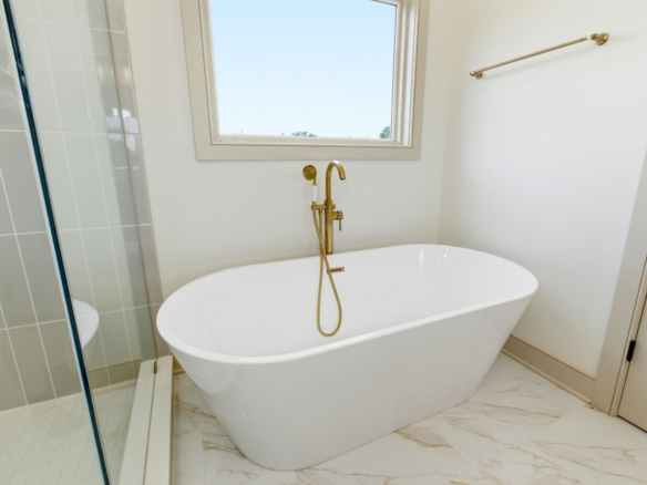 Freestanding white oval tub with gold faucet and handheld shower in a bright bathroom, window above and glass shower to the left.