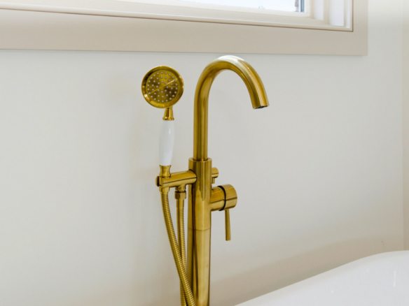 Gold bathroom faucet with a round handheld shower head and flexible hose mounted on a white wall beside a sink.