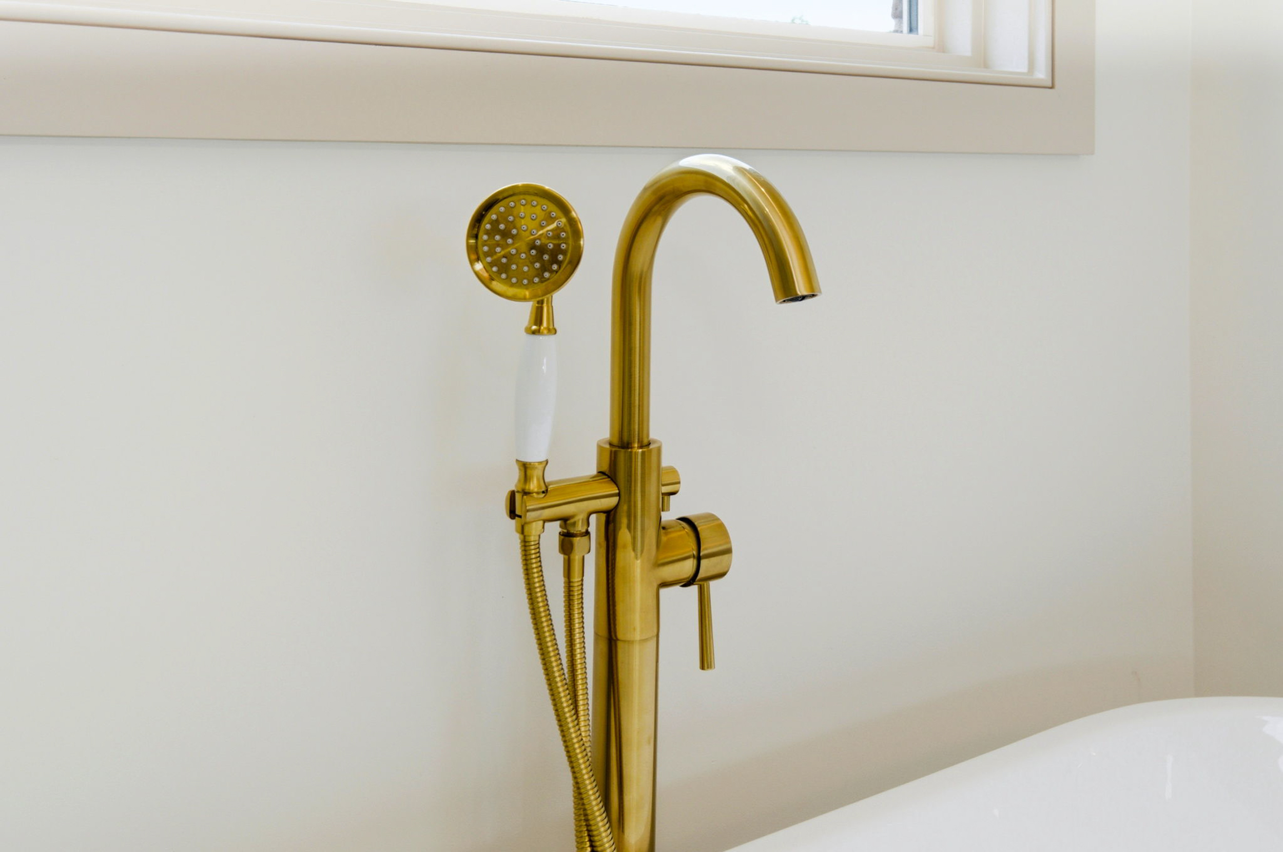 Gold bathroom faucet with a round handheld shower head and flexible hose mounted on a white wall beside a sink.
