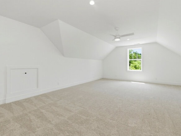 Spacious, bright attic room with white walls, ceiling fan, and window view.