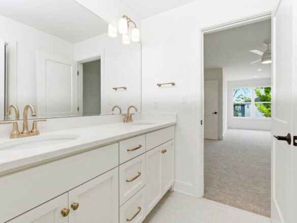 Bright white bathroom with double vanity and gold fixtures, view into bedroom.