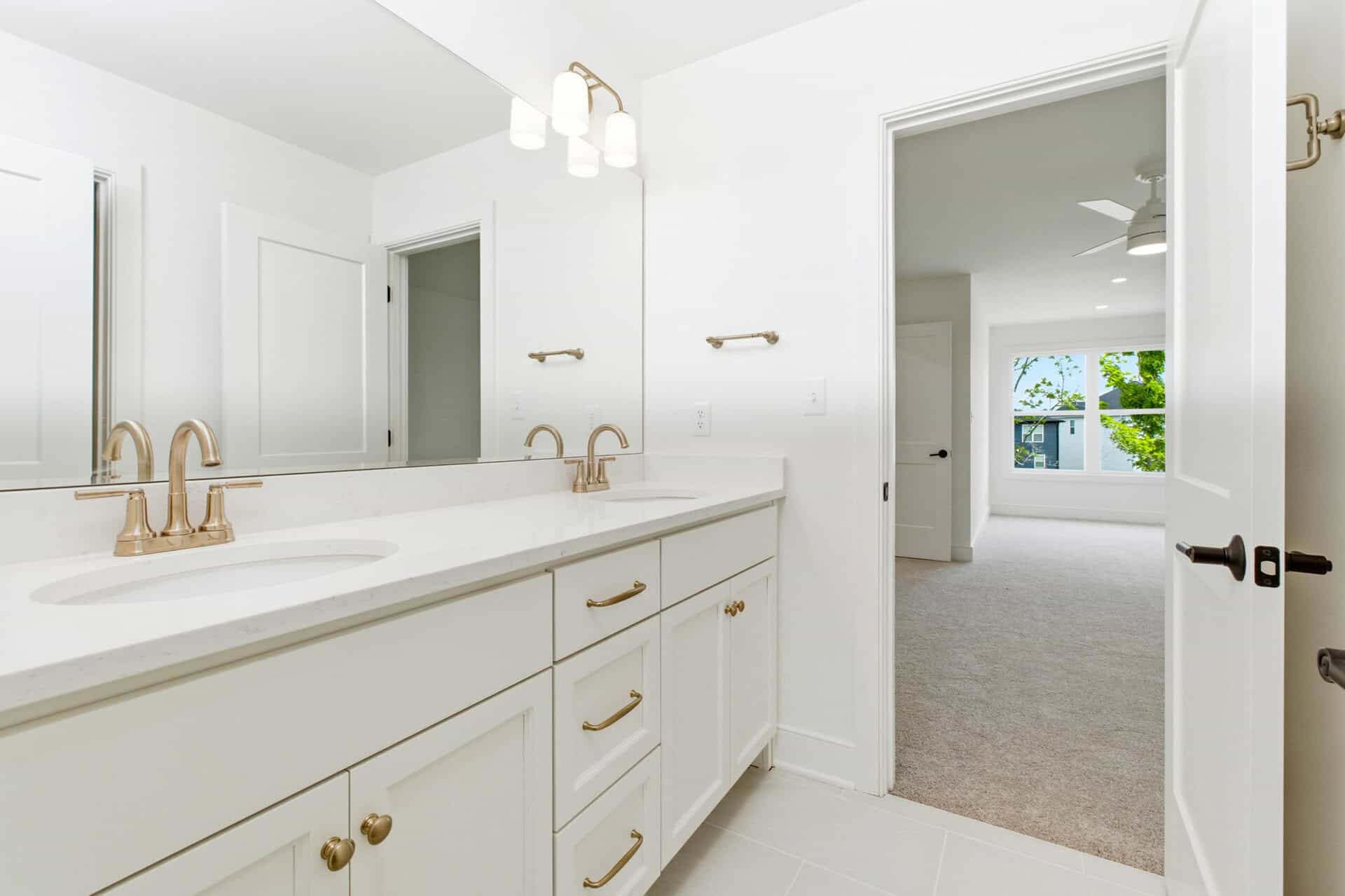Bright white bathroom with double vanity and gold fixtures, view into bedroom.