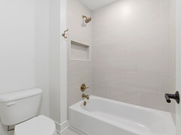 White bathroom with a tub/shower combo, brass fixtures, light gray tile walls, a recessed niche, and a toilet in the foreground.