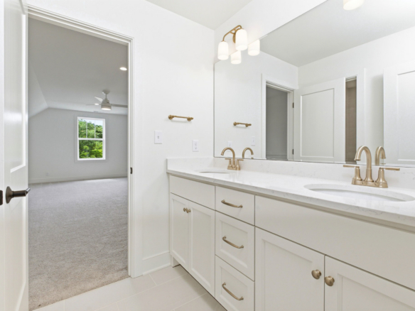 Bright bathroom with a double white vanity, marble countertop, and brass fixtures; large mirror and doorway to a carpeted bedroom with a ceiling fan visible in the adjacent room.