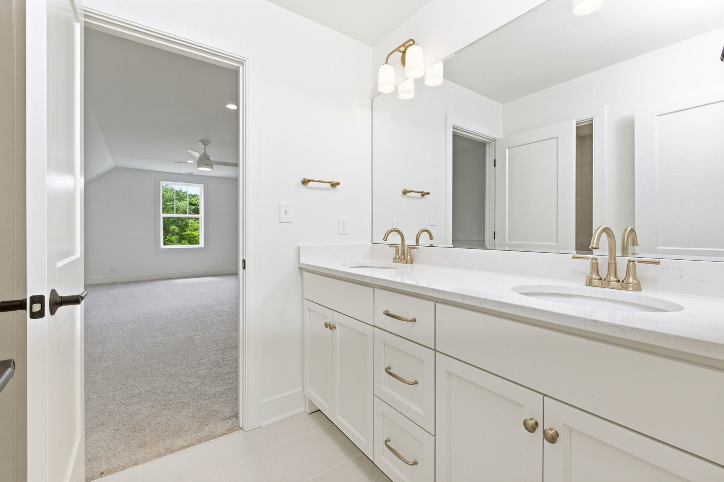 Bright bathroom with a double white vanity, marble countertop, and brass fixtures; large mirror and doorway to a carpeted bedroom with a ceiling fan visible in the adjacent room.