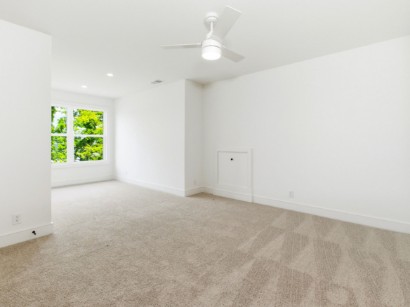 Bright empty room with white walls, beige carpet, a ceiling fan, and a window showing green trees outside.