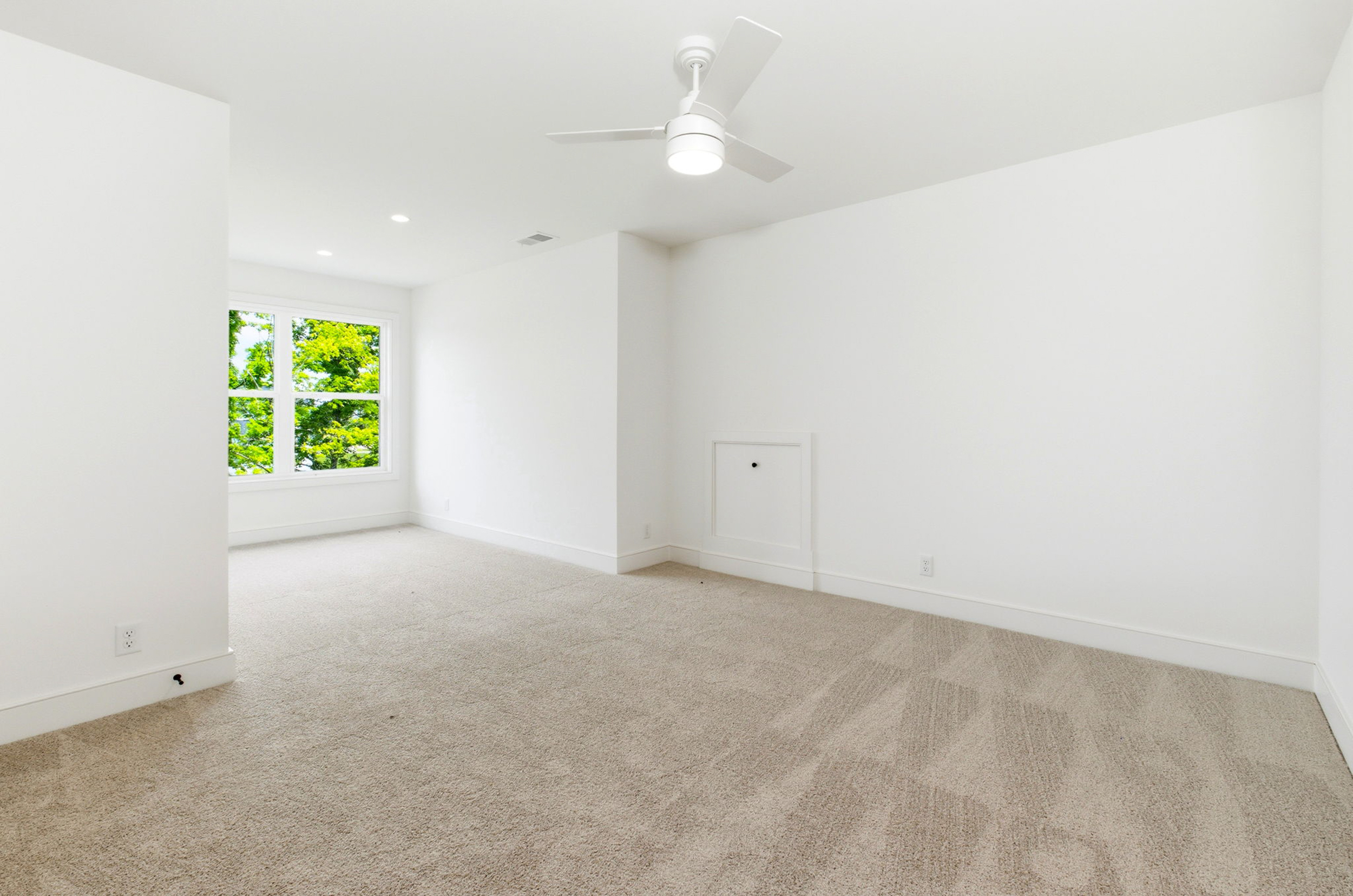 Bright empty room with white walls, beige carpet, a ceiling fan, and a window showing green trees outside.