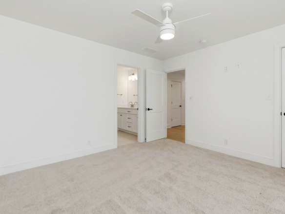 Empty white bedroom with beige carpet, ceiling fan, and two open doors leading to a bathroom and hallway.