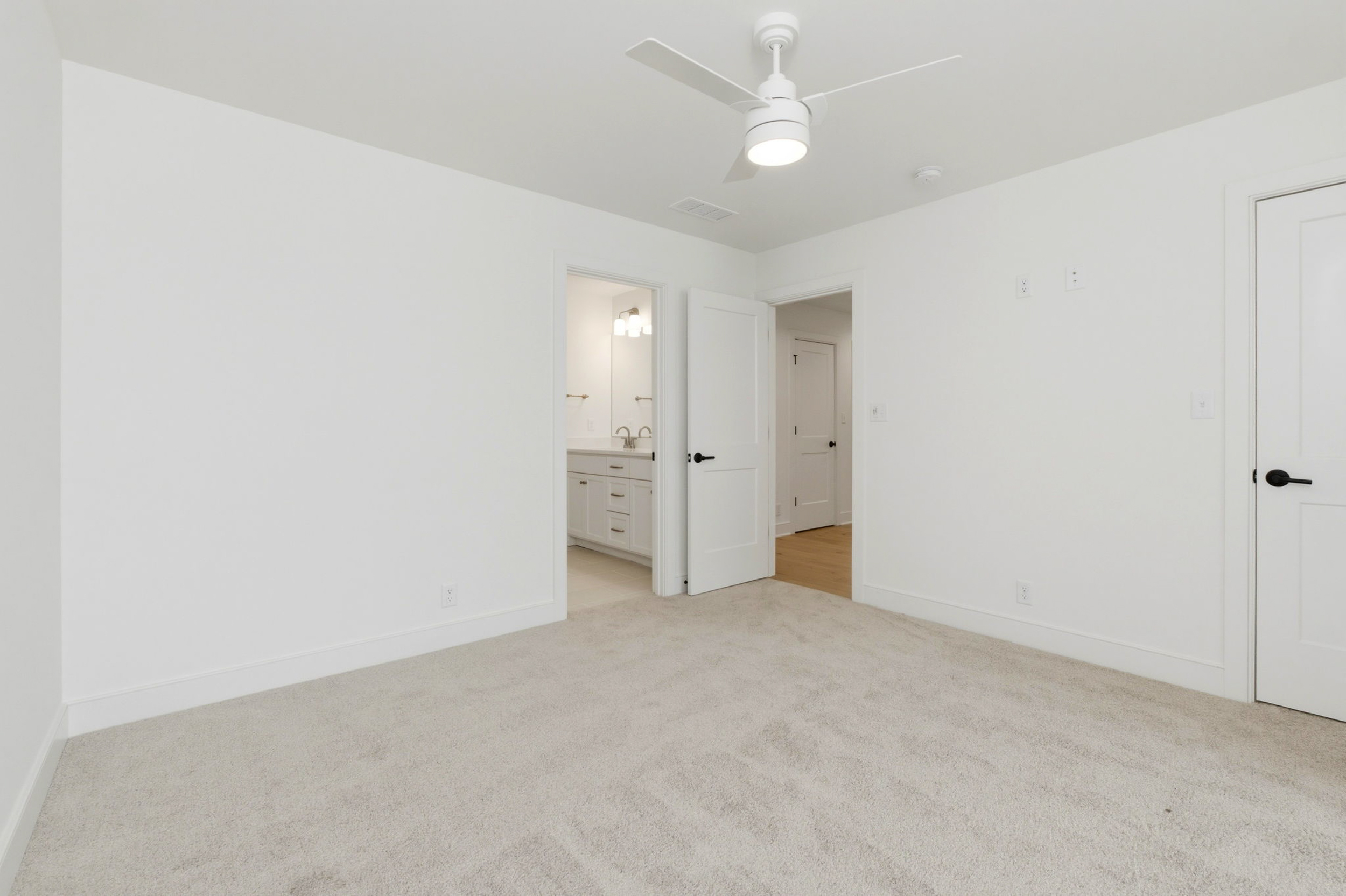 Empty white bedroom with beige carpet, ceiling fan, and two open doors leading to a bathroom and hallway.
