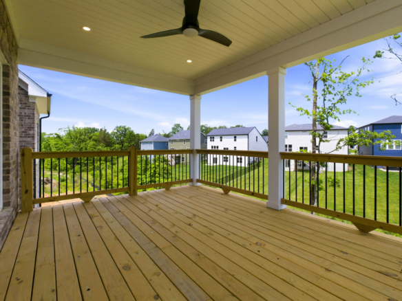 Wooden covered deck with a ceiling fan and black railing, looking out over a green suburban neighborhood under a blue sky.