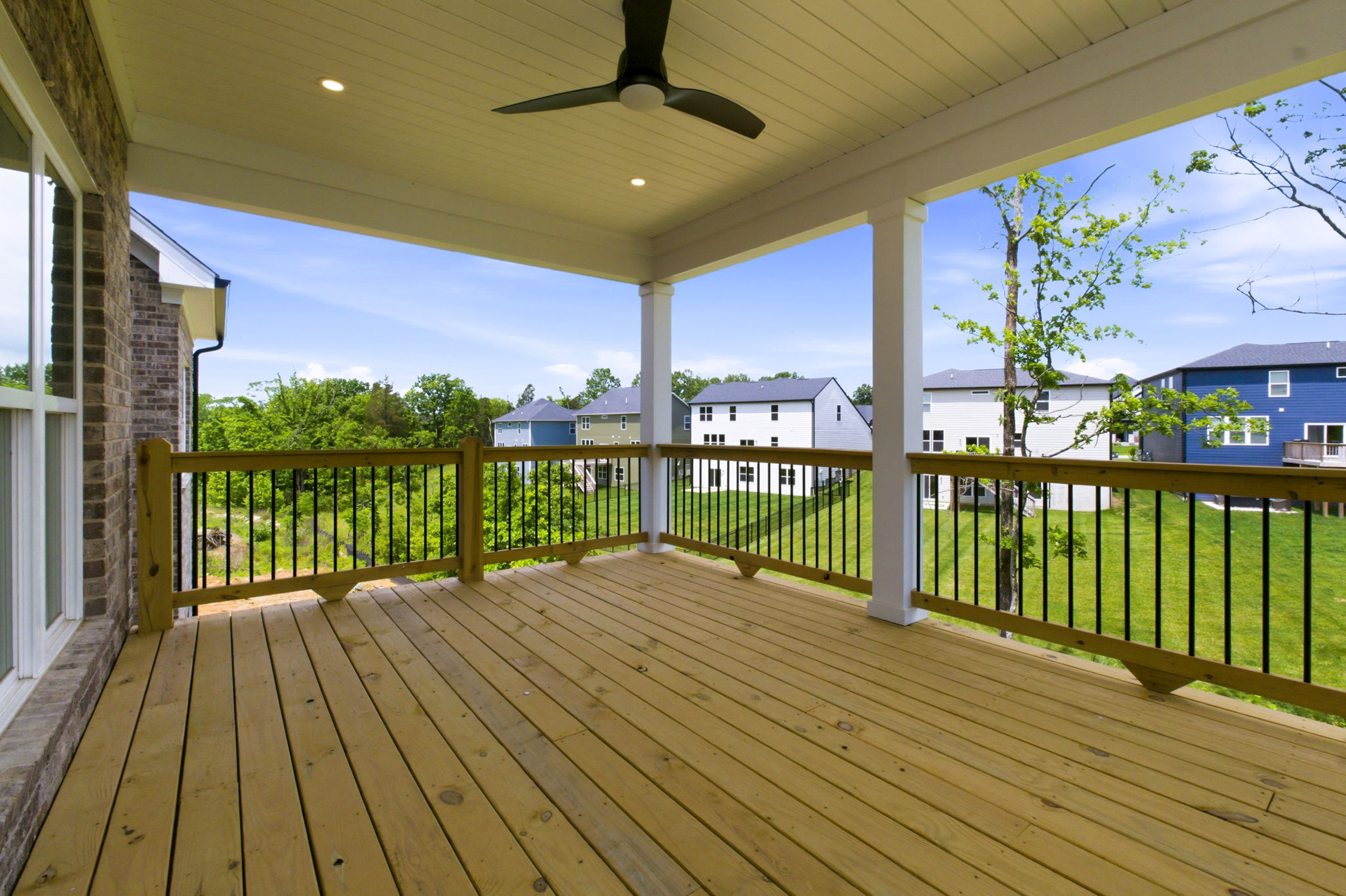 Wooden covered deck with a ceiling fan and black railing, looking out over a green suburban neighborhood under a blue sky.
