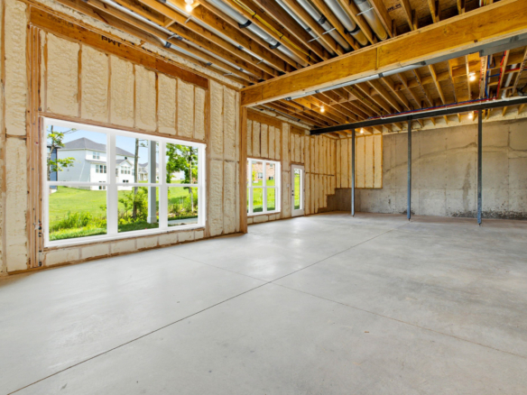 Unfinished interior of a house with foam insulation, wooden studs, exposed ceiling pipes, and large windows letting in daylight.