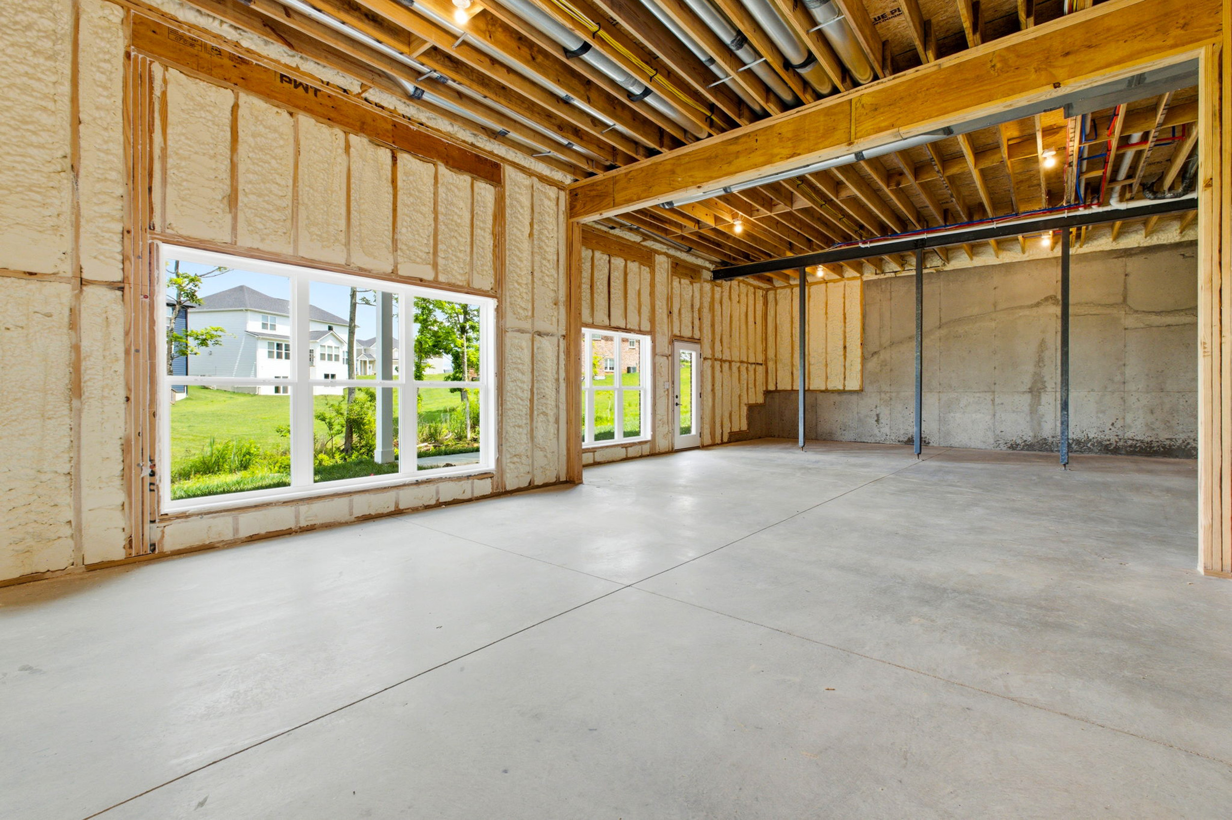 Unfinished interior of a house with foam insulation, wooden studs, exposed ceiling pipes, and large windows letting in daylight.
