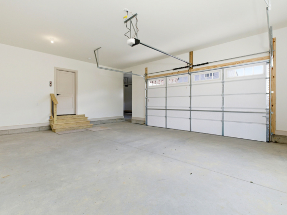 Spacious, empty garage with a side door, wooden stairs, and a large metal sectional door; concrete floor and overhead opener.