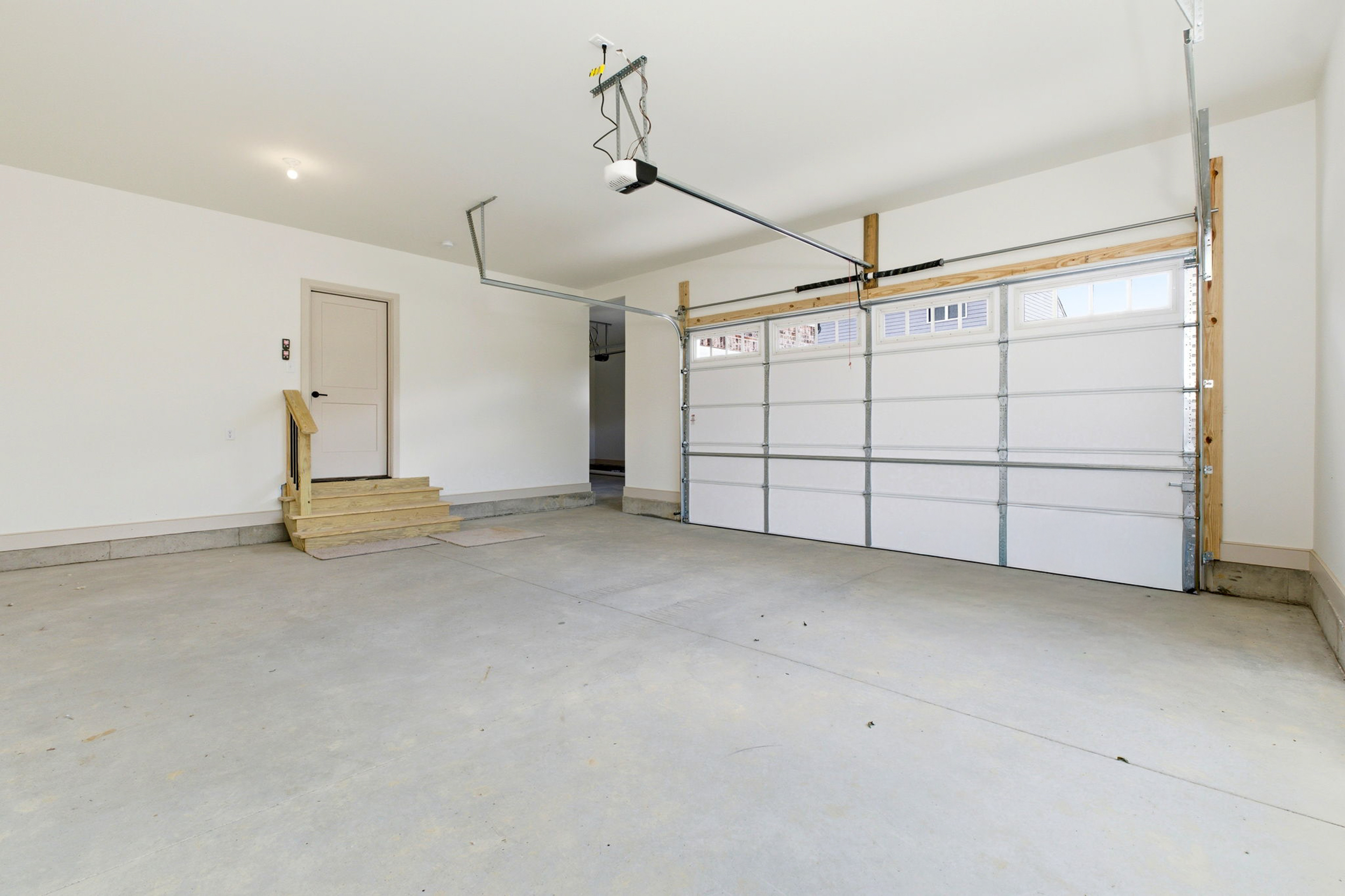 Spacious, empty garage with a side door, wooden stairs, and a large metal sectional door; concrete floor and overhead opener.