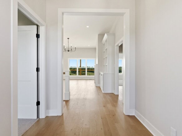 Bright hallway with light wood flooring, white walls and door frames; open doors lead to a sunlit room with a chandelier and large windows showing greenery.